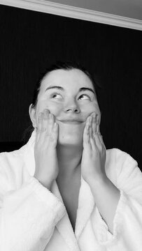 Black and white vertical video. Young adult woman in white bath robe applies moisturizer to face in hotel room. Concept of rest, skin protection during cold season, relax, body care, clean. Wife