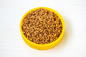 Fenugreek seeds on a yellow container on a white wooden background. Locally in Bangladesh, it is known as Methi Dana.  - obrazy, fototapety, plakaty
