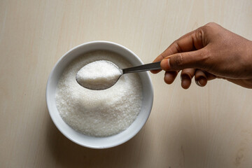 A white bowl is filled with sugar on wooden background. Woman's hand holding a sugar-filled spoon....