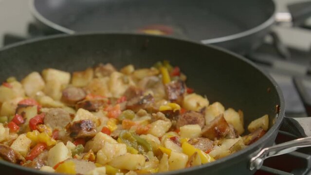 A breakfast hash simmers in a cast iron skillet, with sausages and potatoes, while eggs are cracked into a frying pan.