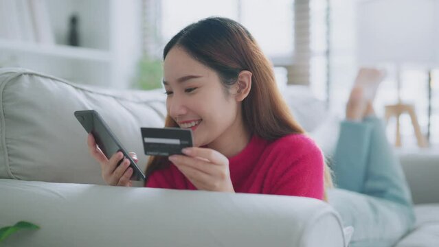 Young asian woman lying on sofa in living room, makes online banking payments through the internet from bank card on cell phone. Shopping online on mobile with credit card