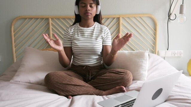 Slow motion video of Black woman doing meditation on bed following online wellness program using laptop and headphones.