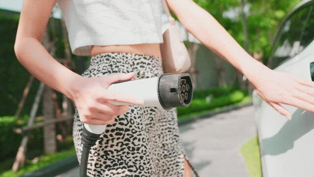 Young woman travel with EV electric car charging in green sustainable city park garden in summer to go shopping in sustainable urban lifestyle with rechargeable electric vehicle innards