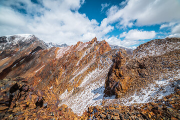 Scenic top view from precipice edge to multicolor big mountain top in freshly fallen snow in sunlight under cloudy sky. Large multi-color snowy rocky ridge. Snow-covered sharp rocks in high mountains.