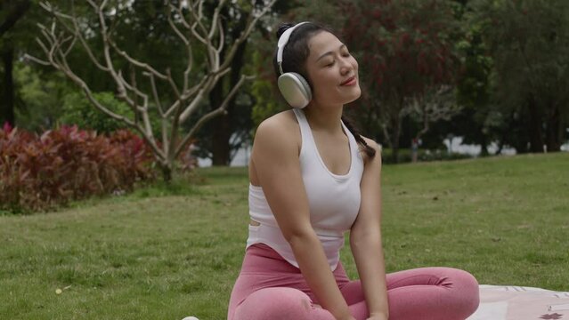Asiatic woman listening music with headphones in park sitting cross legged on yoga mat and wearing sportswear taking break from daily workout