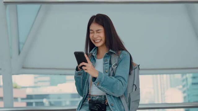 Overjoyed young Asian woman reading message from friend on mobile expressing happiness about great news, emotional female in excited with winning discount for shopping while walking in downtown