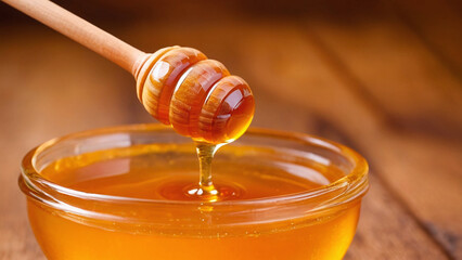 Honey dripping from a wooden honey dipper into a glass bowl - obrazy, fototapety, plakaty