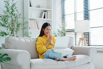Young asian woman in good spirits working on laptop at home while sitting on couch. Excited female using computer notebook, wearing hoodie shirt and happy celebrating success