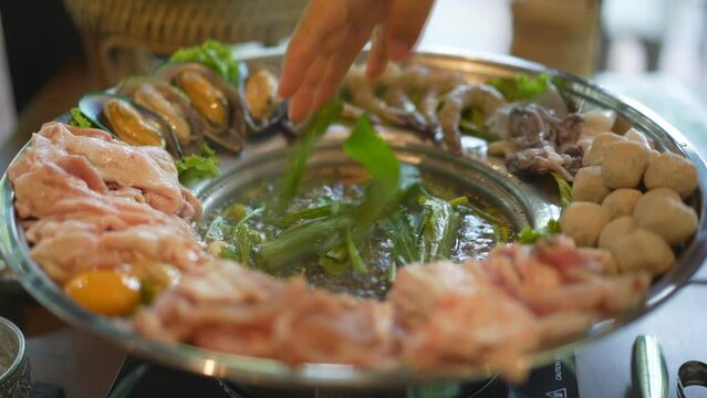 jaew hon shabu or isan hot pot with people hand putting vegetables in soup and raw pork egg meat ball with seafood to boiling spicy water for lunch by thai street food at restaurant on slow motion