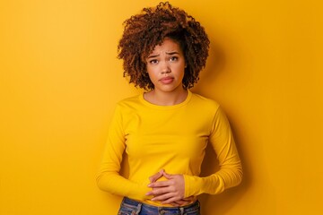 a worried woman in a yellow shirt touching her stomach on yellow studio background