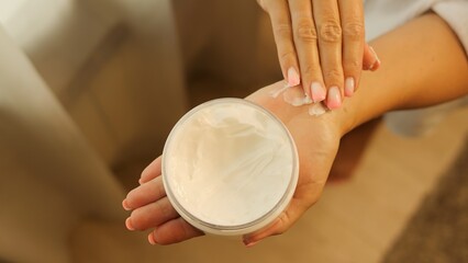 Woman's hand scoops cream from jar, carefully massages onto skin. Cream's application highlights...