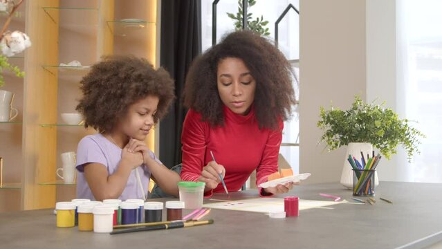 Happy adorable preadolescent girl with curly hair and positive attractive African American mother having fun, blow painting picture with straw on paper, developing child creativity and imagination.