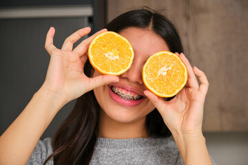 Happy female teenager with braces covering her eyes with an orange fruit.