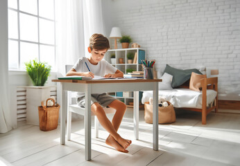 Kind Junge sitzt entspannt in hellem Kinder Jugend Zimmer barfuß am Schreibtisch schreibt lernt...