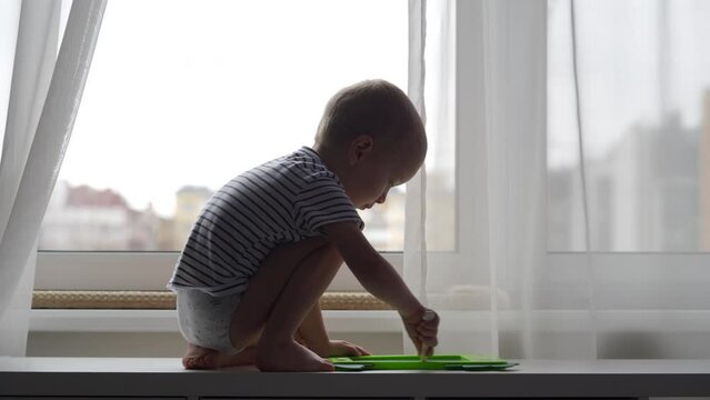 3-year-old child at the window drawing with pen on electronic drawing writing board. Little boy playing with drawing tablet for kids.