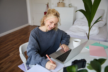 Portrait of smiling, beautiful blond woman, writing down notes, doing homework, studying from home,...