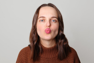 Funny flirting brown haired woman wearing brown jumper standing isolated over light gray background sending air kissing looking at camera with pout lips