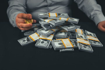 A man spins a Bitcoin coin in his hand over stacks of money in a bank tape Sitting at a table against a black background a man rotates a bitcoin coin and evaluates the money earned on cryptocurrency. - obrazy, fototapety, plakaty