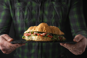 Man wearing checkered shirt and holding freshly baked croissant with iberian ham, tomatoes and salad filling. Close up