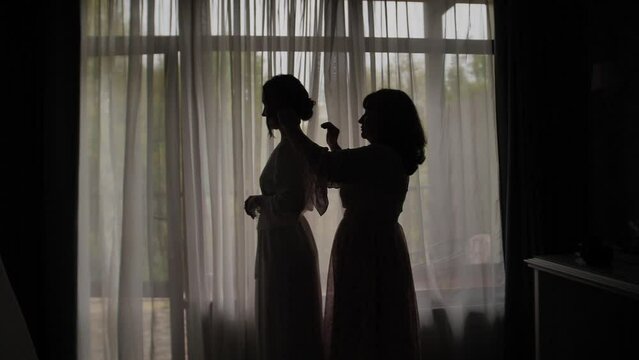 Silhouette of a mother combing her daughter's hair at the window. 
 Silhouette of mother braids her child against a window at sunrise.