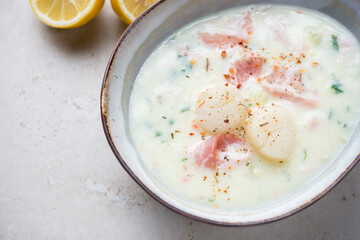 Bowl of creamy chowder with scallops and bacon, horizontal shot on a light-beige stone surface, middle close-up