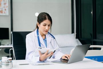 Beautiful physician doctor sit on table in office hospital. Attractive therapist female practitioner pharmacist smile and use tablet and laptop.