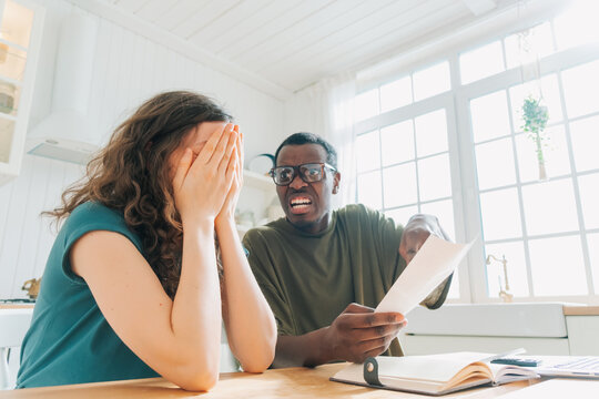 A man is outraged by an expensive check after his wife's purchases. A woman covers her face in dismay while a man looks on with an expression of concern, both reacting to a document in a bright