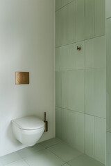 Fototapeta na wymiar A toilet in the bathroom, pale green tiles on the wall, brass sink and rectangular light grey tile with a square opening for a hanger and shelf, white floor, minimalist interior design.