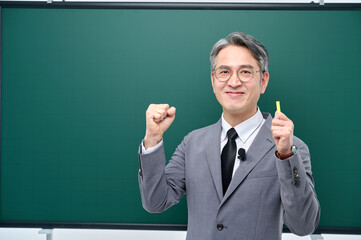 A middle-aged male teacher in a suit and glasses is giving an Internet lecture with a confident expression and pose with chalk in front of a green blackboard.
