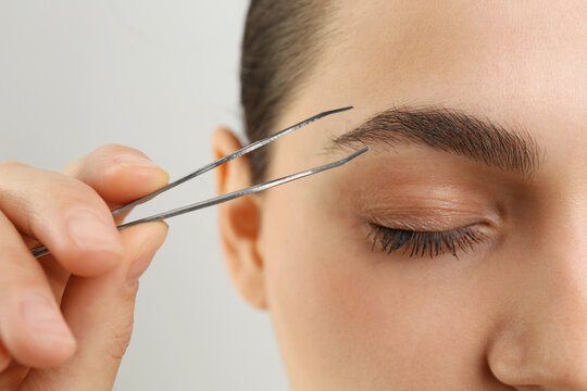 Eyebrow correction. Young woman with tweezers on light grey background, closeup