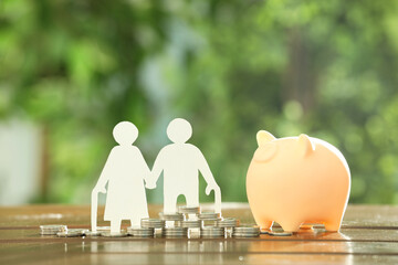 Pension savings. Figure of senior couple, piggy bank and coins on wooden table outdoors