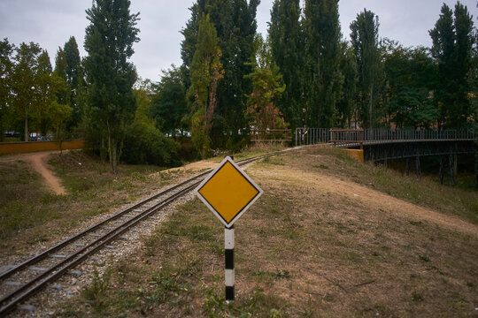 Yellow train signal with train tracks on the right at sunset and a defocused railway bridge surrounded by vegetation