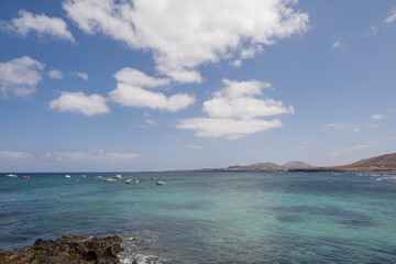 Seascape. Rocks in the foreground. Group of boats anchored nearby. Mountains in the background. Turquoise Atlantic Ocean. Big white clouds. Village of Arrieta. Lanzarote, Canary Islands, Spain