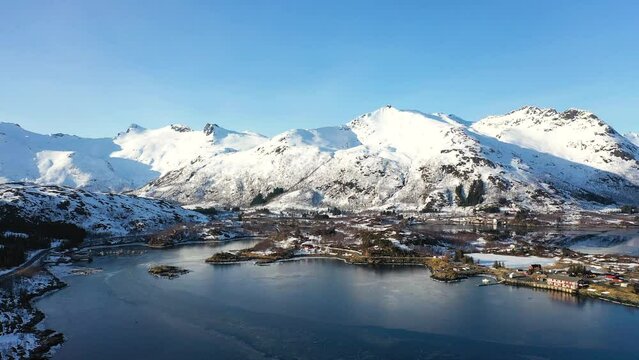 Incredible travel. Aerial view from flying drone of sea shore, snowy mountains, rorbuer in winter at sunset. Lofoten islands, Norway. Top drone view of fjord, village, reflection in water, rocks.