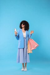 Happy young woman with shopping bags and modern smartphone on light blue background