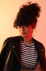 Beautiful young woman in black leather jacket posing on color background in neon lights