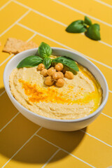 A dish of hummus with chickpeas and basil on a yellow tile table