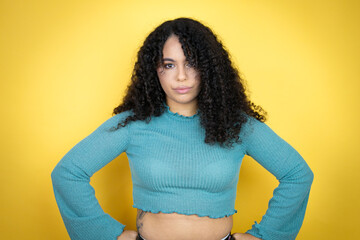 African american woman wearing casual sweater over yellow background skeptic and nervous, disapproving expression on face with arms in waist - obrazy, fototapety, plakaty