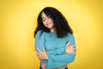 African american woman wearing casual sweater over yellow background hugging oneself happy and positive, smiling confident - obrazy, fototapety, plakaty