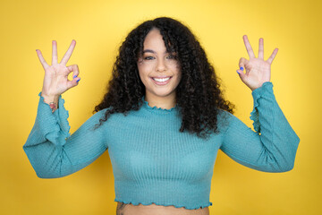 African american woman wearing casual sweater over yellow background doing ok sign with fingers and...