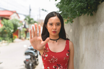 Young Filipino woman in a red dress, gesturing stop on the sidewalk, walking in a lower middle...