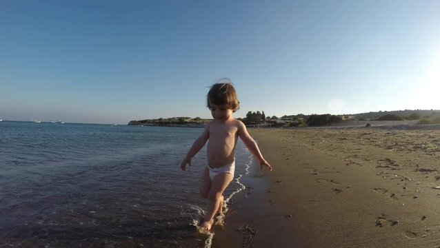 Toddler baby girl in nappy running on sandy beach through sea waves
