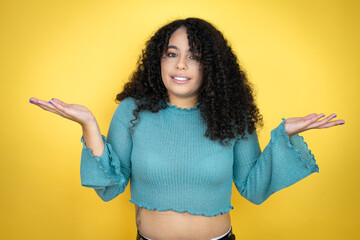 African american woman wearing casual sweater over yellow background clueless and confused expression with arms and hands raised - obrazy, fototapety, plakaty