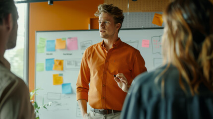 A man in an orange shirt is gesturing during a team meeting with a whiteboard in the background. - obrazy, fototapety, plakaty