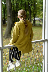 A girl with a long braid in white sneakers sits on an openwork white fence of a gazebo with her...