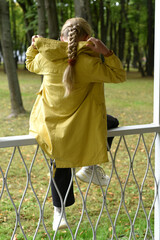 A girl with a long braid in white sneakers sits on an openwork white fence of the gazebo with her...