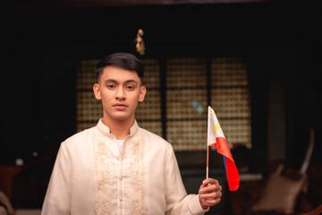 A serious and patriotic young FIlipino man in traditional Barong Tagalog shirt and slacks, and holding a Philippine flag, inside an ancestral house.