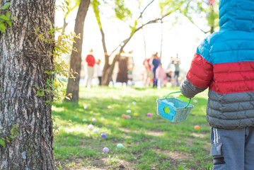 Asian kid in sweatpants, jacket with hood holding Easter basket looking down green grass meadow with colorful eggs ready for egg hunt tradition at local Church in Dallas, Texas, decorated Paschal