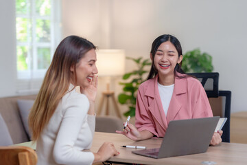 Two asian woman colleagues share a joyful moment of laughter, showcasing a positive work relationship in a bright home office.