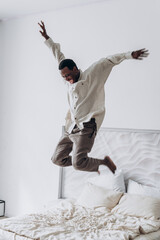 Exuberant joy radiates from a man as he leaps into the air above a bed, a spontaneous moment captured against a clean, white backdrop. Happy African American man jumps on the bed